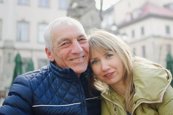 Hombre mayor guapo está abrazando a su joven esposa rubia pasar tiempo juntos al aire libre en la ciudad antigua durante principios de primavera u otoño . — Foto de Stock