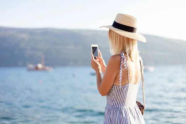 Woman traveler is using mobile phone at summer sea beach. Female tourist is taking photos and smiling. Girl in hat and casual clothes is traveling and enjoying vacation.