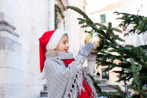 Kid is decorating Christmas tree with golden ornaments outdoors. Happy child is enjoying holidays.