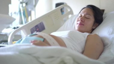 Hamile Asyalı Çinli kadın hastane yatağında uzanıyor ve doktoru bekliyor.
