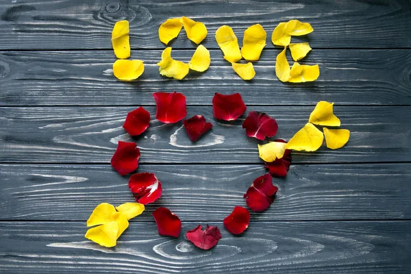 Heart lined with red petals and pierced with an arrow with white flowers. The heart is labeled with yellow petals word love. Gray wooden background