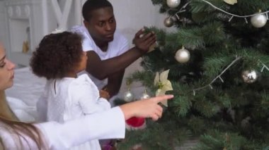 Noel ağacında sallanan mutlu bir kız bebek. Noel ağacını süsleyen çocuklar. Yılbaşı ve yeni yıl ağacının arka planında beyazlar içinde mutlu, çok ırklı bir aile.