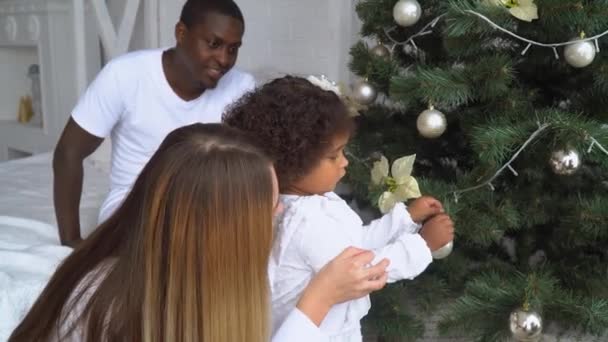 Joyeux bébé fille suspendue boule sur un arbre de Noël. Des enfants décorent un sapin de Noël. Jeune heureuse famille multi-ethnique vêtue de blanc à l'arrière-plan de Noël et Nouvel An arbre à la maison — Video