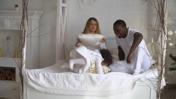 Happy mixed ethnicity family mom, african dad and little kid daughter having fun pillow fight on bed. Young multi-ethnic family dressed in white at background of Christmas tree in home — Stock Video