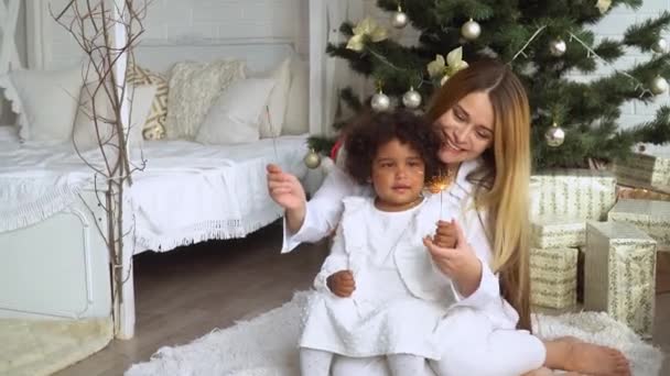 Madre feliz con hija pequeña vestida de blanco sentada junto al árbol de Navidad y sosteniendo chispa — Vídeos de Stock