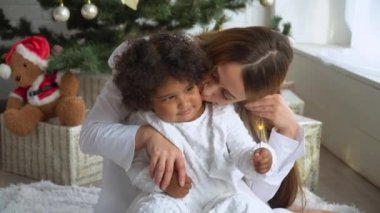 Beyaz elbiseli mutlu bir anne Noel ağacının yanında oturmuş maytap tutuyor.