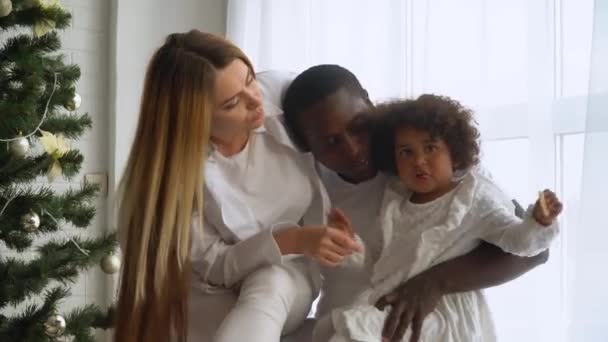 Jeune heureuse famille multi-ethnique de mère, père et petite fille vêtus de blanc assis près du sapin de Noël. bébé fille manger biscuits — Video