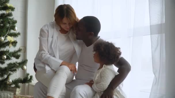 Молода щаслива багатоетнічна сім'я матері, батька і маленької дочки, одягнена в біле, сидячи біля ялинки — стокове відео