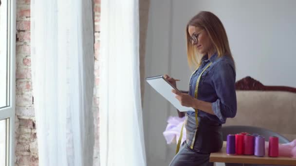 Linda costureira loira fica ao lado da janela com uma fita métrica em torno de seu pescoço e um caderno em suas mãos e faz anotações — Vídeo de Stock