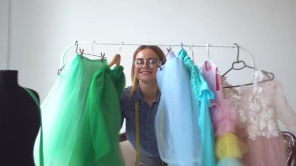 Mooie blonde naaister kijkt vrolijk uit van achter kleren op een hanger — Stockvideo