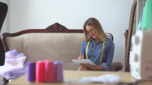 Mooie naaister blond in een naaiatelier zit op een bank en schetst haar nieuwe idee in een notebook — Stockvideo