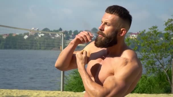 Bodybuilder fume une cigarette. Jeune bodybuilder athlétique barbu fait du sport en plein air . — Video