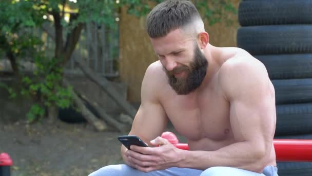 Bodybuilder utiliza smartphone. Joven culturista atlético barbudo practica deportes al aire libre — Vídeos de Stock