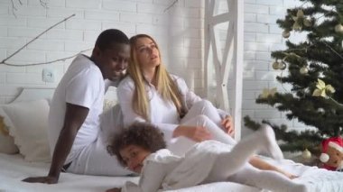Evde Noel ve yılbaşı ağacının arka planında beyaz giyinmiş anne, baba ve küçük kızın mutlu çok ırklı aileleri var. Çok ırklı aile, Xmas konsepti.