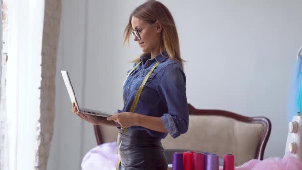 Linda loira costureira fica ao lado de uma janela aberta com uma fita métrica em torno de seu pescoço e trabalha em um netbook — Vídeo de Stock