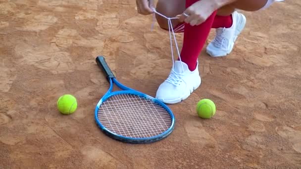 Egy teniszező cipőfűzőt köt a teniszpályára. Egy ütő és két teniszlabda fekszik a pályán. — Stock videók