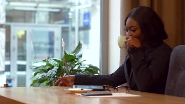 Αφροαμερικανή επιχειρηματίας που δουλεύει με λάπτοπ και χαρτιά. Κορίτσι που πίνει καφέ — Αρχείο Βίντεο