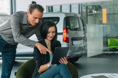 Woman and man wearing smart casual wear in auto show on car background and working with tablet. Consultant at a car dealership presenting a car for a woman of buyers