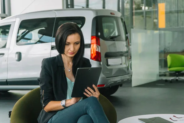 Una mujer con una ropa casual inteligente se sienta en un espectáculo de coches en el fondo de un coche y trabaja con una tableta — Foto de Stock