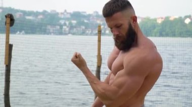 Vücut geliştirici kaslarını gösterir. Genç sakallı atletik vücut geliştirici dışarıda spor yapıyor.