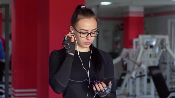 Mladá dívka se sluchátky používající chytrý telefon v tělocvičně. Dívka poslouchá hudbu — Stock video