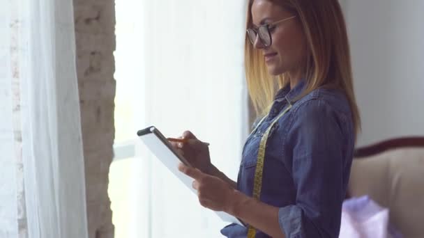 Beautiful blonde seamstress stands by the window with a meter tape around her neck and a notebook in her hands and makes notes — Stock Video