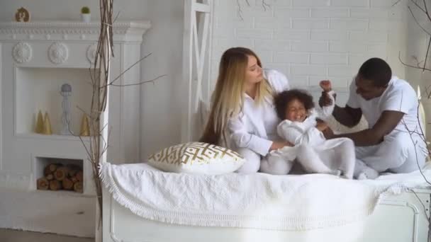 Joven feliz familia multiétnica de la madre, el padre africano y la pequeña hija vestida de blanco está sentado en la cama en casa — Vídeos de Stock