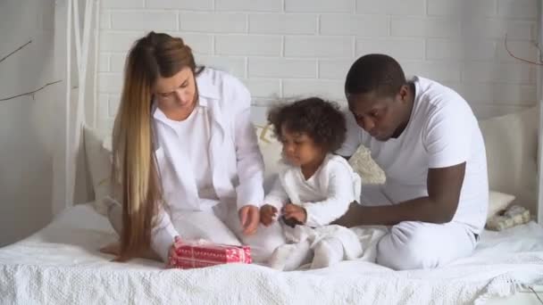 Молодая счастливая многонациональная семья матери, африканский отец и маленькая дочь, одетые в белое сидит на кровати в доме — стоковое видео
