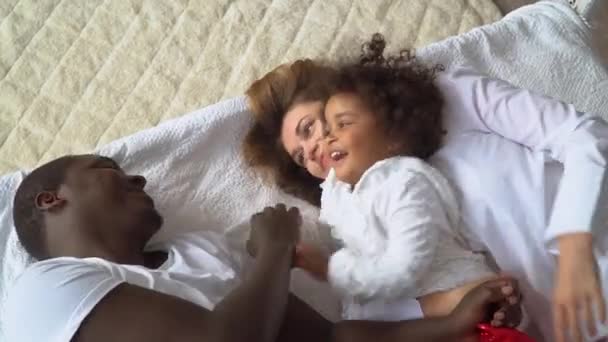 Jovem família multi-étnica feliz de mãe, pai africano e filha vestida de branco está deitada na cama em casa — Vídeo de Stock