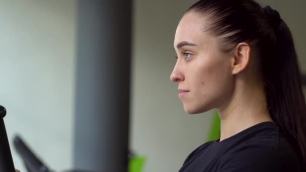 Retrato de una joven hermosa en el gimnasio en una bicicleta estacionaria — Vídeos de Stock