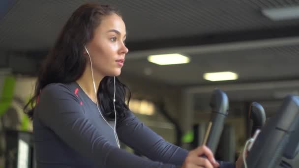Retrato de uma jovem menina bonita com fones de ouvido no ginásio em uma bicicleta estacionária — Vídeo de Stock