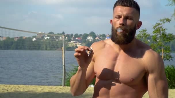 Bodybuilder smokes a cigarette. Young bearded athletic bodybuilder plays sports outdoors — Stock Video