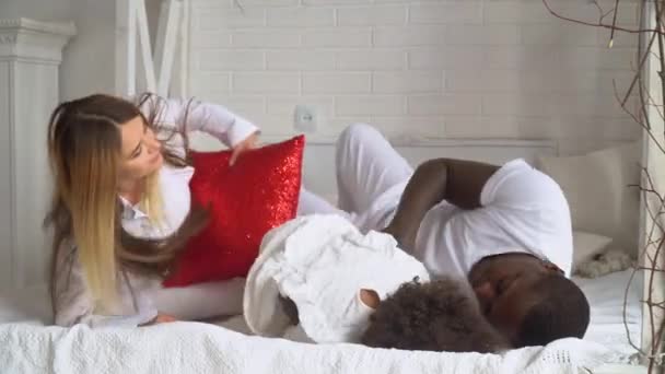 年轻快乐的多民族家庭，母亲、非洲裔父亲和身穿白衣的小女儿躺在家里的床上 — 图库视频影像