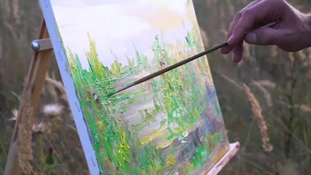 Artista masculino pinta paisaje al atardecer en el espacio abierto en medio del paisaje natural. Vista de cerca de los movimientos de lienzo y pincel en una mano — Vídeos de Stock