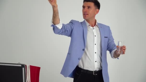 Magician in a suit shows a trick with glass and water on a white background — Stok video