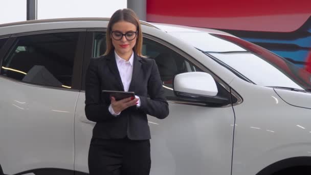 La vendeuse se tient près de la voiture dans le concessionnaire automobile et prend des notes sur la tablette — Video