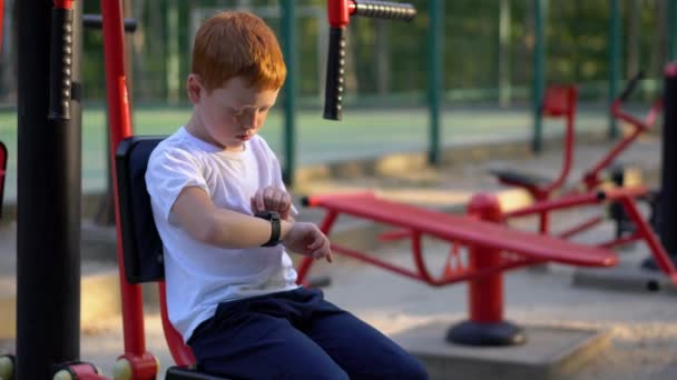 A boy on street public exercise equipment looks at his wristwatch — Stok video