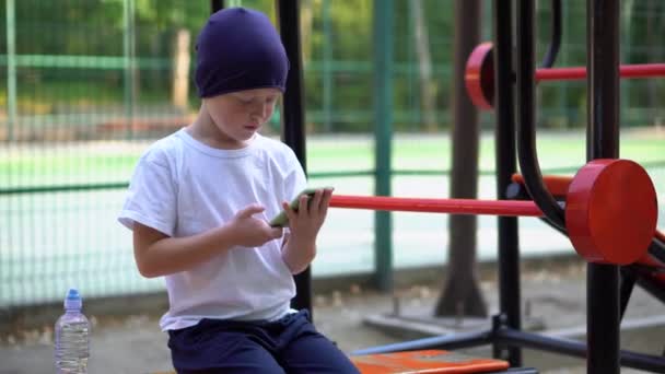 路上で公共の運動機器の少年は彼のスマートフォンを見て — ストック動画