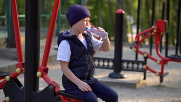 屋外の公共フィットネス機器の近くの小さな男の子飲料水。子供スポーツ — ストック動画