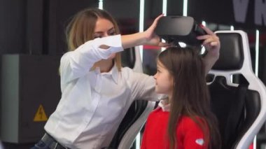 Bir kadın kızına gözlük takar. Kadın ve küçük kız sanal gerçeklik simülatörü oynamaya hazırlanıyorlar.