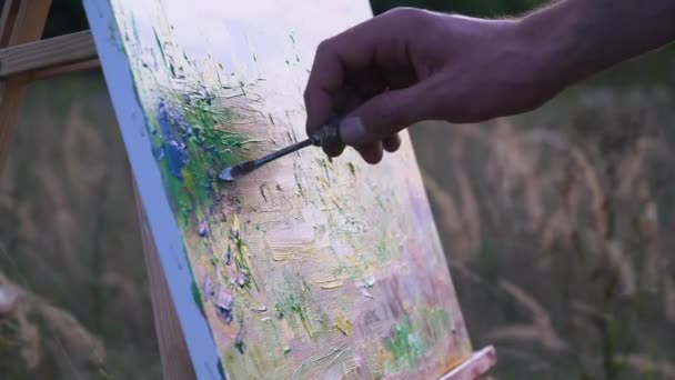 Мужчина рисует пейзаж заката в открытом пространстве посреди природного ландшафта. Крупный план движения холста и кисти в руке — стоковое видео