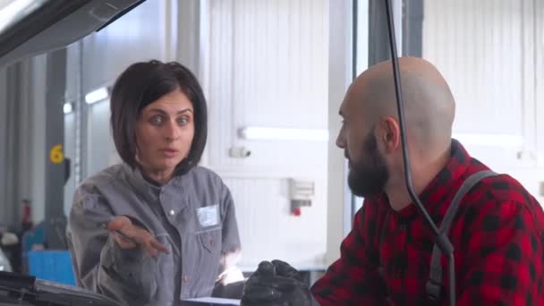 Dos mecánicos profesionales hembra y macho reparando un coche en el taller de reparación de automóviles. La jefa regaña a su subordinado — Vídeos de Stock