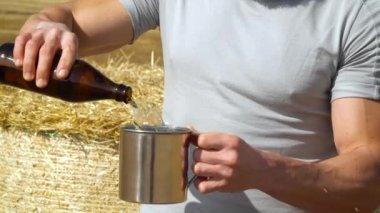 Bir adam buğday tarlasında balyalı metal bir bardağa bira döker. Taze bira, güneş enerjisi