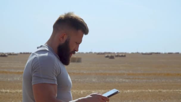 Granjero barbudo sostiene una tableta en sus manos sobre un fondo de fardos de heno — Vídeos de Stock