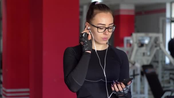 Mladá dívka se sluchátky používající chytrý telefon v tělocvičně. Dívka poslouchá hudbu — Stock video