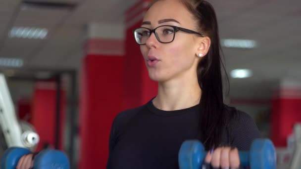 青いダンベルでジムで運動する若いフィットネス女性。スポーツクラブのメガネの美しい女の子は、シミュレータ上の演習の異なる種類を行います — ストック動画