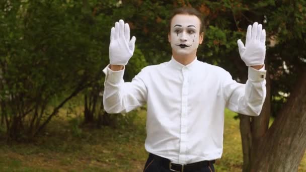 Jeune mime drôle derrière une boîte en verre invisible ou un mur. Mime sur le parc montre pantomime — Video