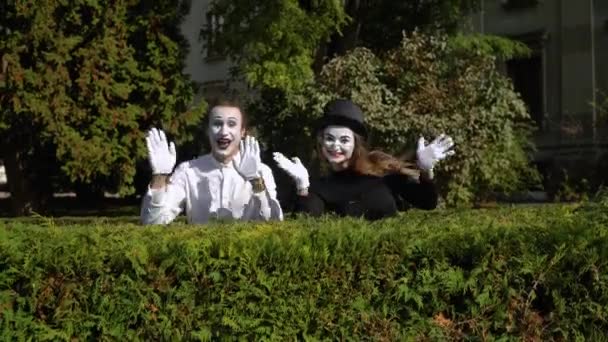 Zwei Pantomimen im Park, die sich hinter Büschen verstecken. Mime und Mimin schauen in die Kamera und lächeln auf grünem Hintergrund in einem Park — Stockvideo