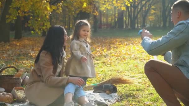 Familia soplando burbujas de aire. Madre y padre con hija divirtiéndose al aire libre. Picnic en el parque de otoño — Vídeos de Stock