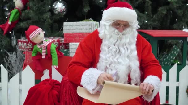 De Kerstman zit met een zak cadeautjes op het keizerrijk en schrijft iets op de lijst. — Stockvideo
